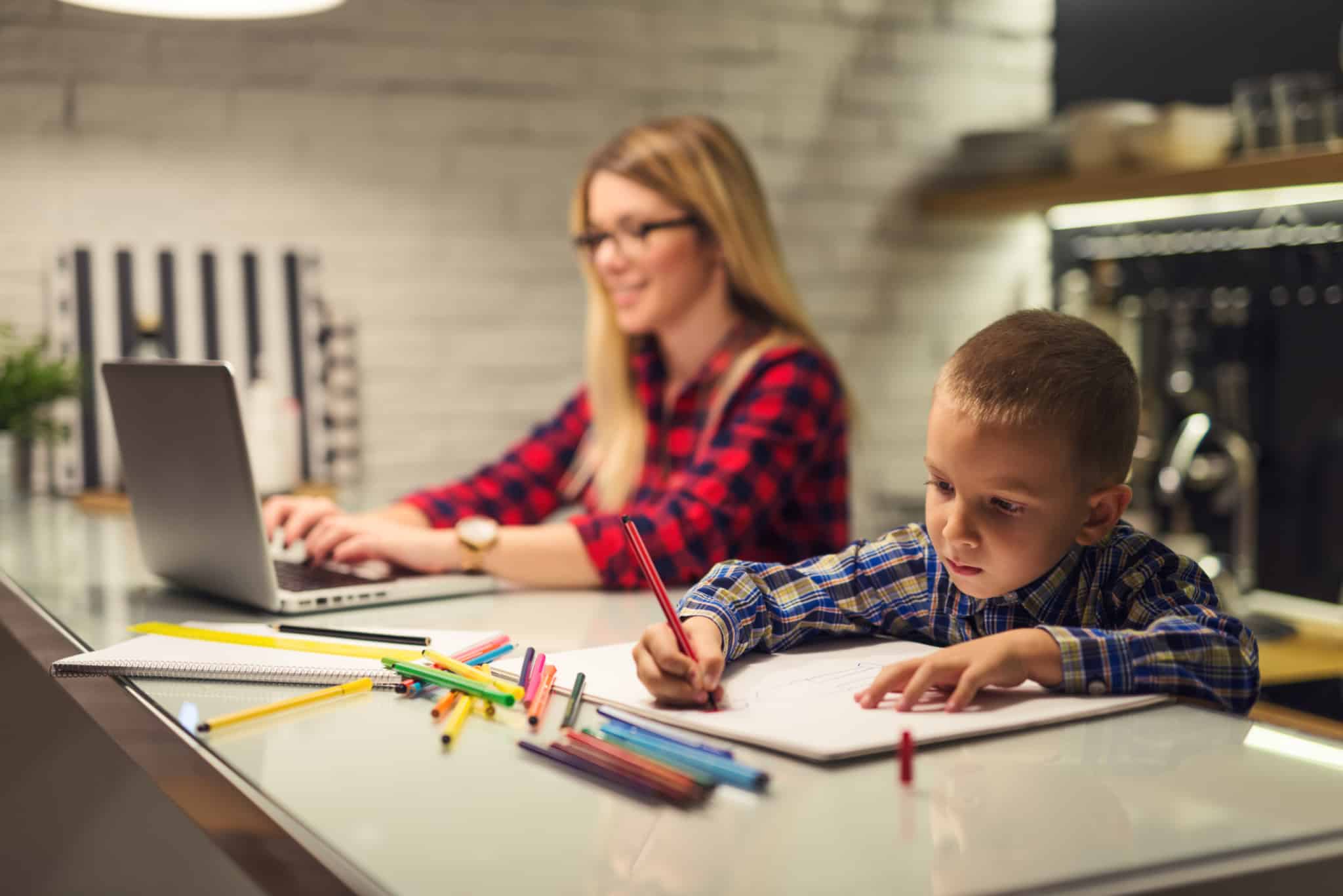 Female working on laptop with little boy coloring on paper next to her